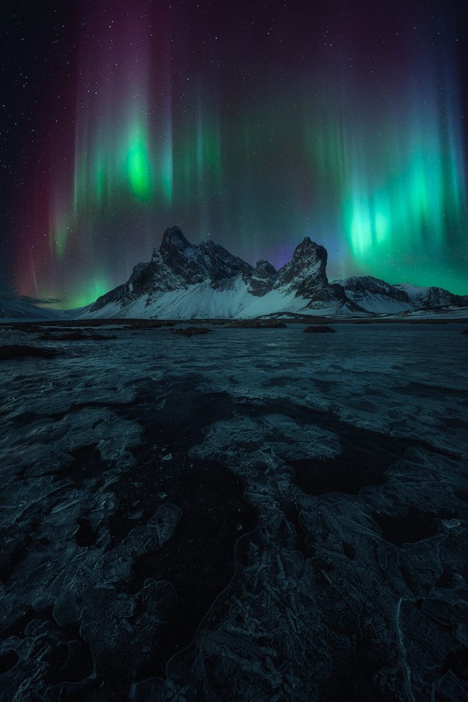 Night photo of an icy lake, with mountains and northern lights in the sky in the background, with stars glimpsed through